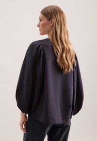 Kvinde med langt blondt hår iført en løs sort bluse med flagrende ærmer og mørke jeans, vender sidelæns mod en neutral baggrund.