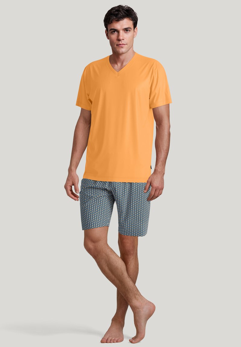 Oranje V-hals t-shirt van soepele stof, gecombineerd met gestreepte blauwe shorts met kleine geometrische vormen. Model staat op blote voeten.