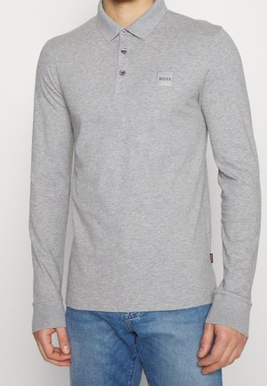 Mann trägt ein hellgraues Langarm-Polo mit „BOSS“-Logo auf der Brust, kombiniert mit blauen Jeans vor einfachem Hintergrund.