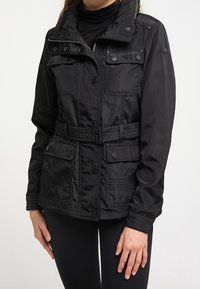 Veste noire légère avec fermeture éclair et boutons-pression, plusieurs poches, taille ceinturée et col montant. Texture lisse.