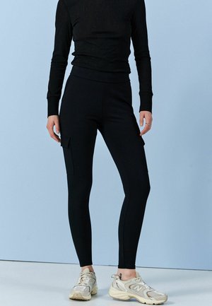 Henkilö, joka on pukeutunut mustiin korkeavyötäröisiin cargo-leggingseihin ja valkoisiin lenkkareihin, seisoo vaaleansinistä taustaa vasten.