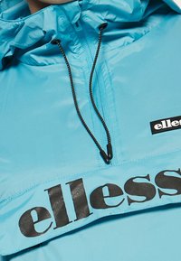 Světle modrá pulovrová bunda s vysokým límcem, zapínáním na zip a černým logem na hrudi, vyrobená z hladkého, voděodolného materiálu.