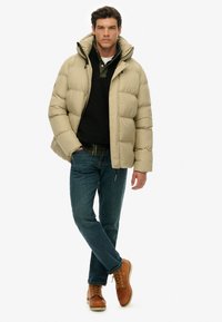 Beige puffjakke med hætte, lynlås fortil og elastiske mansjetter over en mørk sweater og en ternet skjorte. Bæres med blå jeans og brune støvler.