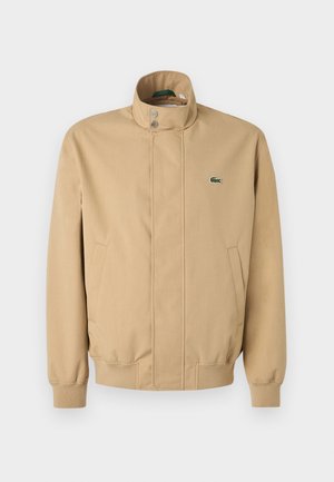 Giacca beige con colletto alto, bottoni a pressione, tasche laterali e polsini a costine. Presenta un logo Lacoste verde sul petto. Tessuto liscio.