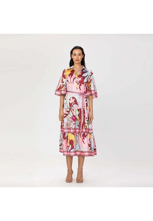 Femme debout portant une robe midi colorée à motifs avec de larges manches et une taille ceinturée, associée à des talons nus à bout ouvert.