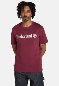Mörkröd bomull t-shirt med Timberland-logotyp och trädgrafik i vitt och rött. Korta ärmar, rund hals och en avslappnad passform.