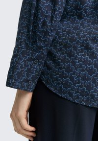 Marineblauwe shirt met een gestructureerd patroon van golvende lijnen in lichtblauw en metallic accenten. Voorzien van een knoopsluiting bij de manchetten met donkere knopen.