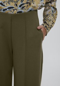Pantalons vert olive, ajustés et taille haute, avec des poches latérales et des plis visibles à l'avant, assortis à une chemise à motif floral.