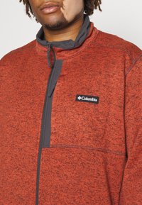 Giacca in pile Columbia di un rosso melange, con chiusura a zip nera, accenti grigi e un piccolo logo sul petto.