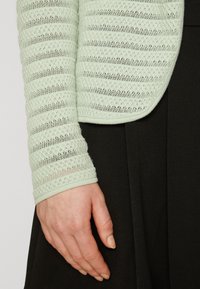 Světle zelený texturovaný kardigan se pruhovaným vzorem a žebrovanými manžetami, kombinovaný s jednobarevným černým kouskem. Zaměření na rukáv a ruku.