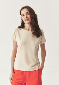 Tricou bej cu mânecă scurtă, cu decolteu larg și mâneci răsucite, asortat cu pantaloni roșii strălucitori cu șnur. Textură netedă, design casual.