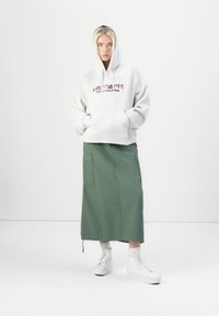 Gray hoodie with a front pocket and red text, paired with a green, ankle-length skirt featuring side pockets. White platform sneakers.
