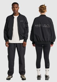 Czarna kurtka bomber z bocznymi kieszeniami, ściągaczami i zapięciem na zamek. Z przodu i z tyłu widnieje szare logo "HALO".