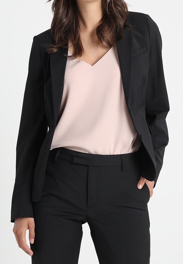 Svart skräddarsydd kavaj med slag, bärs över en ljusrosa silkeblusa. Kavajen har en enda bröstficka och raka byxor.