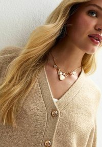 Cardigan beige lavorato a maglia con scollo a V, bottoni dettagliati e una collana in oro con più ciondoli a forma di cuore e perle.