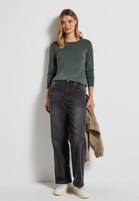 Donkergrijze longsleeve shirt gecombineerd met losse zwarte jeans en witte sneakers; model houdt een lichtgewicht beige jas vast.