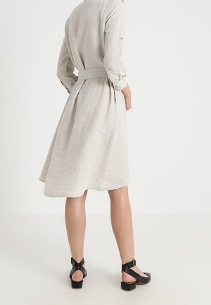 Frau trägt ein knielanges, beiges Leinenkleid mit 3/4-Ärmeln und schwarzen, niedrig-absatz-ankle-strap Schuhen, steht mit dem Rücken zur Kamera vor einem weißen Hintergrund.