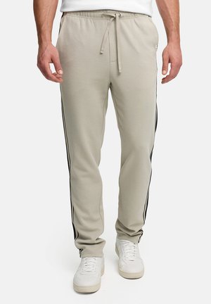 Mand iført beige joggebukser med sorte sidelister og hvide sneakers, stående mod en ensfarvet baggrund.
