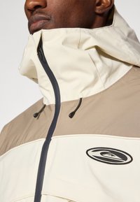 Homem a usar uma jaqueta bege e creme com gola alta e fecho preto, close-up mostrando o emblema no peito.