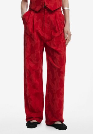 Pantalones rojos de talle alto y pierna ancha con un sutil estampado floral, combinados con zapatos planos negros y un brazalete plateado en un brazo.