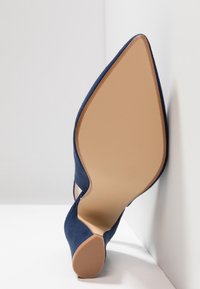Chaussure à talon haut en daim bleu à bout pointu vue par le dessous, mettant en valeur la semelle beige lisse et l'embout du talon texturé.