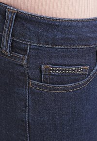 Tmavě modré džíny se strukturovaným povrchem, s zadní kapsou zdobenou černými korálky a kontrastním prošíváním.