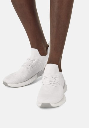 Zapatos atléticos blancos con un upper de punto texturizado, cuello similar a un calcetín y una suela de goma gris. Presenta cordones y acentos de diseño sutiles.
