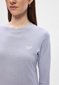 Γυναίκα που φοράει ένα ανοιχτό μωβ μακρυμάνικο πουκάμισο με το λογότυπο "tommy jeans" κεντημένο στο στήθος και με έναν χρυσό κρίκο αυτιού ορατό.