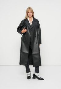 Svart faux lær dobbeltspent frakk med store revers, lange ermer og en jevn tekstur. Brukt over mørke jeans og svarte sko.