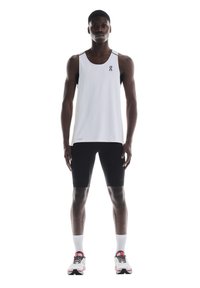 Hvid atletisk tanktop med sorte accenter, parret med sorte tætsiddende shorts. Modellen bærer hvide sokker og sporty sko med røde detaljer.