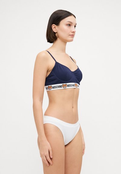 Navy blue bralette with adjustable straps and a contrasting Moschino logo band; paired with white cotton briefs. Smooth, form-fitting design.
