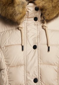 Beige gewatteerde jas met een met bont afgewerkte capuchon, zwarte knopen en een decoratieve trekkoord. Soepele textuur met een doorstikt patroon. 