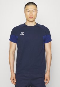 Mężczyzna o krótkich włosach, ubrany w granatowo-niebieską sportową koszulkę hummel z krótkim rękawem, stojący na jednolitym jasnym tle.