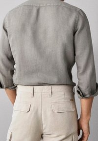 Man in een opgerolde grijze lange mouw shirt, ingestopt in beige broek met achterzakken, getoond van achteren.
