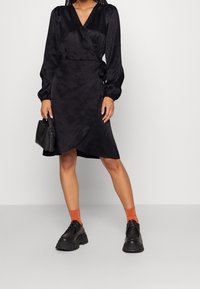 Černé obtažené šaty s dlouhými rukávy, texturovaný materiál a výstřih do V, doplněné chunky černými botami a oranžov�ými ponožkami. Součástí je také černá kabelka.