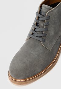 Szare zamszowe botki za kostkę z okrągłym noskiem, sznurowanym designem, kontrastującym brązowym przeszyciem środkowej podeszwy oraz gładką teksturą.