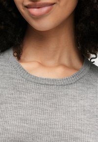 Κοντινές λήψεις του κάτω μέρους του προσώπου και του λαιμού ενός ατόμου που φοράει γκρι ριχτό πουλόβερ με στρογγυλή λαιμόκοψη και έχει σγουρά σκούρα μαλλιά.