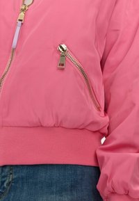Růžová bomber bunda s kapsou na zip, žebrovaným pasem a manžetami. Vyrobena z hladké látky s lesklým povrchem.