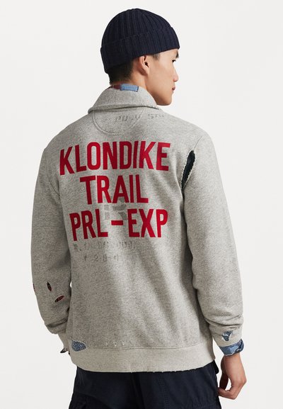 Sivi dukserica s crvenim natpisom "KLONDIKE TRAIL PRL-EXP", sa istrošenim detaljima i mornarsko plavim pletenim šeširom. Američki radnički stil.