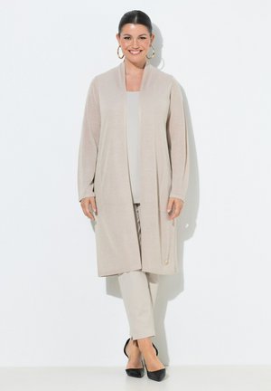 Donna sorridente, in piedi su sfondo bianco, indossa un lungo cardigan beige, top e pantaloni coordinati, décolleté nere con punta a punta e orecchini a cerchio dorati.