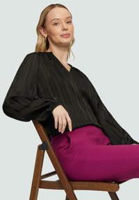Blouse noire à manches longues avec des rayures verticales et un décolleté en V ; associé à un pantalon rose vif ; modèle assis sur une chaise en bois.