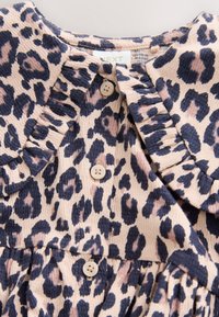 Blouse imprimé léopard avec col à volants et boutons beiges sur tissu texturé, avec des taches noires et marron sur fond clair.