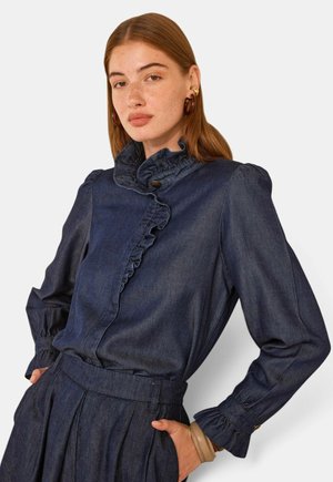 Femme aux cheveux roux portant une blouse en denim foncé avec un col et des poignets volantés, associée à un pantalon en denim taille haute assorti, mains dans les poches.