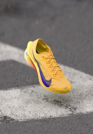 Zapatilla de running amarilla de Nike con swoosh púrpura suspendida en el aire sobre un asfalto gris con una marca de flecha blanca.