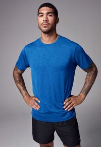 Uomo con tatuaggi che indossa una maglietta sportiva blu e pantaloncini neri, in piedi con le mani sui fianchi davanti a uno sfondo grigio.