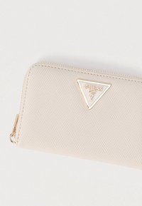 Portefeuille zippé texturé beige clair avec un logo triangulaire Guess USA bordé d'or et une tirette de fermeture éclair dorée.