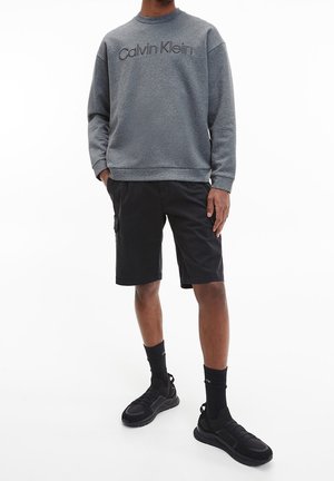 Sudadera gris con el logo de "Calvin Klein", combinada con pantalones cortos negros y zapatillas deportivas negras. Modelo con calcetines negros en una pose de pie.