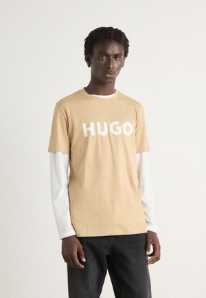 Lichtbruine t-shirt met "HUGO" in het wit erop gedrukt. Langere witte mouwen zijn zichtbaar eronder, gecombineerd met donkere jeans. Gladde katoen textuur.