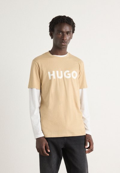 Светлокафяв тишърт с бяло изписано "HUGO". Дълги бели ръкави са видими отдолу, съчетани с тъмни дънки. Гладка памучна текстура.