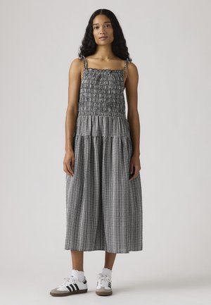 Mujer de pie, vestida con un vestido midi de cuadros vichy en blanco y negro, cuerpo fruncido y tirantes con lazos en los hombros, calcetines blancos hasta el tobillo y zapatillas blancas.